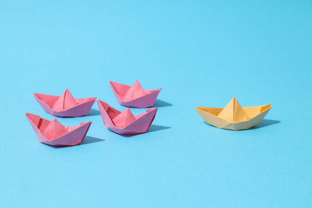 Five paper boats with one different colored in front of the four